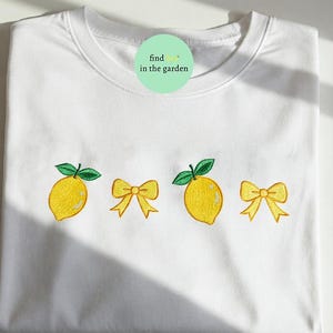 Puede incluir: Sudadera blanca de cuello redondo con bordados de limones y lazos amarillos. El dise&ntilde;o incluye dos limones con hojas verdes y dos lazos amarillos. Un c&iacute;rculo verde con el texto "find in the garden" est&aacute; cerca del cuello.