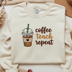 May include: Cream-colored sweatshirt with the embroidered text "coffee teach repeat" next to an embroidered coffee cup with a heart. The coffee cup has a brown cup, a black heart, and a green straw.