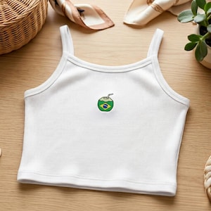 Pode incluir: Top curto branco canelado com alças finas. O top apresenta um pequeno desenho bordado de um coco com canudo e a bandeira brasileira. O top é feito de um material macio e confortável.