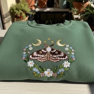 May include: A green sweatshirt featuring an embroidered moth design. The moth is brown and white, surrounded by a floral wreath with crescent moons and stars. The sweatshirt is folded neatly on a wooden surface.