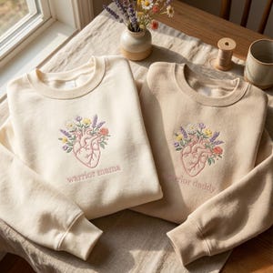 Peut inclure: Deux sweat-shirts crème avec des motifs de cœur floral brodés. L'un dit "warrior mama" et l'autre "warrior daddy". Les sweat-shirts sont faits d'une matière douce, idéale pour les tenues décontractées.