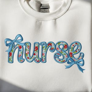 Könnte beinhalten: Weißes Sweatshirt mit dem Wort "nurse", das in einem Blumenmuster mit roten und blauen Blumen gestickt ist. Das Wort ist blau umrandet und hat blaue Schleifen.