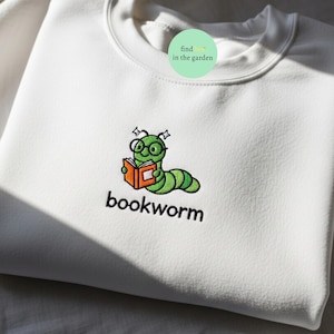 May include: White crew neck sweatshirt with an embroidered design. The design features a green bookworm wearing glasses and reading an orange book, with the word "bookworm" embroidered below.