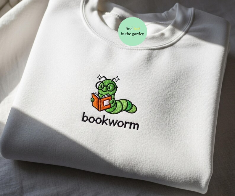 May include: White crew neck sweatshirt with an embroidered design. The design features a green bookworm wearing glasses and reading an orange book, with the word "bookworm" embroidered below.
