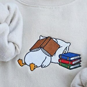 May include: Cream-colored sweatshirt with an embroidered design of a cartoon character sleeping with a book over its face. A stack of books in blue, red, and green are next to the character. The sweatshirt's sleeves are visible.