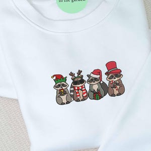 May include: White sweatshirt featuring an embroidered design of four raccoons dressed in festive holiday attire. Each raccoon wears a different hat, including an elf hat, reindeer antlers, a Santa hat, and a top hat. They are holding candy canes and gifts.