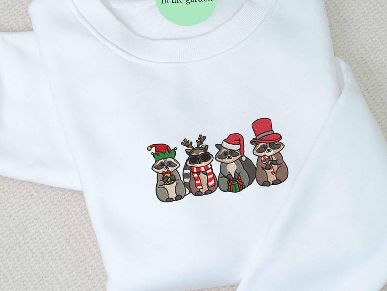May include: White sweatshirt featuring an embroidered design of four raccoons dressed in festive holiday attire. Each raccoon wears a different hat, including an elf hat, reindeer antlers, a Santa hat, and a top hat. They are holding candy canes and gifts.