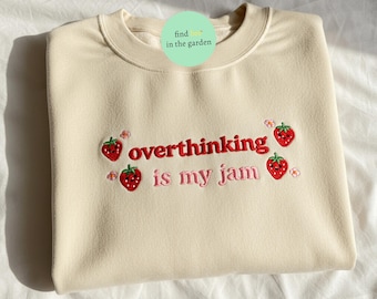May include: Cream-colored sweatshirt with the embroidered phrase "overthinking is my jam" in red and pink. The design includes embroidered strawberries and small flowers. The sweatshirt has a crew neck and long sleeves.