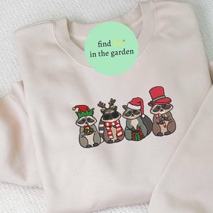 May include: A cream-colored sweatshirt featuring an embroidered design of four raccoons dressed in festive holiday attire. Each raccoon wears a different hat and holds a candy cane or gift. The text "find me in the garden" is on the top.