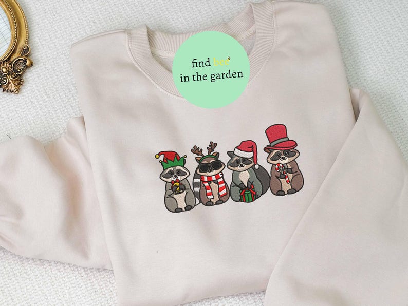 May include: A cream-colored sweatshirt featuring an embroidered design of four raccoons dressed in festive holiday attire. Each raccoon wears a different hat and holds a candy cane or gift. The text "find me in the garden" is on the top.