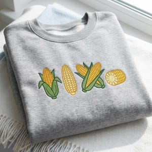 May include: A gray sweatshirt featuring an embroidered design of corn on the cob. The design includes four corn illustrations: two whole ears, one partially husked, and a cross-section. The sweatshirt is folded on a white blanket.