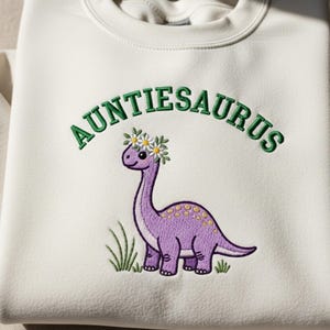 May include: Cream-coloured sweatshirt featuring the embroidered text "AUNTIESAURUS" in green above a purple dinosaur wearing a flower crown. The dinosaur has yellow spots and stands in front of green grass.