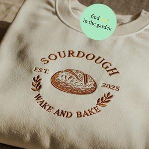 Può includere: Una felpa beige con un logo ricamato marrone che dice "SOURDOUGH EST. 2025 WAKE AND BAKE". Il logo presenta un pane.