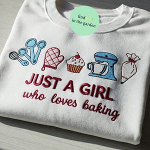 Può includere: Felpa bianca con ricamo rosso che dice "Just a girl who loves baking". Il ricamo include un cupcake, una planetaria, una sac à poche, una frusta e un guanto da forno rosso.