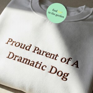 May include: White sweatshirt with brown embroidered text that reads "Proud Parent of A Dramatic Dog".