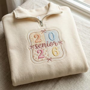 Könnte beinhalten: Cremefarbenes Sweatshirt mit einem Viertel-Reißverschluss-Ausschnitt. Auf der Vorderseite befindet sich ein gesticktes Emblem mit dem Text "2026 Senior" in Pastellfarben, verziert mit Blumen- und Schleifendetails. Das Sweatshirt ist ordentlich gefaltet.