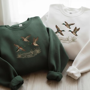 May include: Two sweatshirts, one forest green and one white, are displayed. Each features an embroidered design of flying ducks above a stylized marsh scene. The sweatshirts have long sleeves and a crew neck.