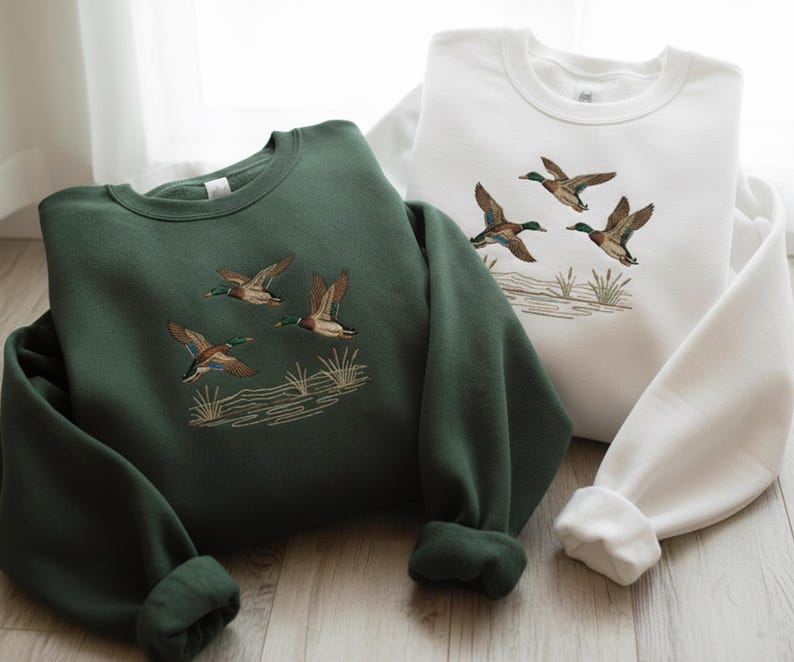 May include: Two sweatshirts, one forest green and one white, are displayed. Each features an embroidered design of flying ducks above a stylized marsh scene. The sweatshirts have long sleeves and a crew neck.