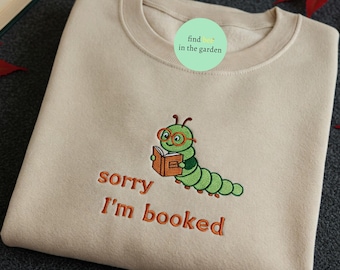 May include: A beige sweatshirt with a green worm wearing glasses and reading a book. The text "sorry I'm booked" is embroidered in orange below the worm.