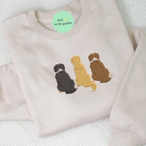 Peut inclure: Un sweat-shirt beige avec un cercle vert qui dit "find in the garden" et trois chiens brodés en noir, marron et jaune.