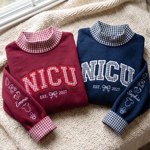 Op de afbeelding: Twee sweatshirts met lange mouwen in bordeauxrood en marineblauw, elk met een opstaande kraag en boorden met een vichy-ruitpatroon. De voorzijde toont de tekst NICU, EST. 2027 en een strik. De mouwen bevatten geborduurde baby-iconen en de namen Emma en Sophia.