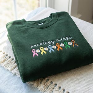 May include: A dark green sweatshirt featuring the embroidered words "oncology nurse" in white. Below the text are embroidered ribbons and daisies in various colours: pink, yellow, light blue, orange, green, and rainbow.
