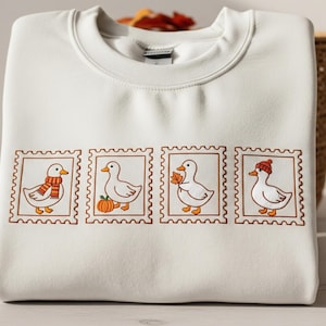 Könnte beinhalten: Cremefarbenes Sweatshirt mit vier gestickten Briefmarkenmotiven mit Enten. Jede Ente ist mit herbstlichen Accessoires geschmückt: einem Schal, einem Kürbis, einem Blatt und einem Hut. Das Sweatshirt ist gefaltet.