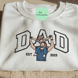 May include: White sweatshirt with the word "DAD" in a black outlined collegiate font. An illustration of a father holding a child on his shoulders is centered below the word. The text "EST 2025" is below the illustration.