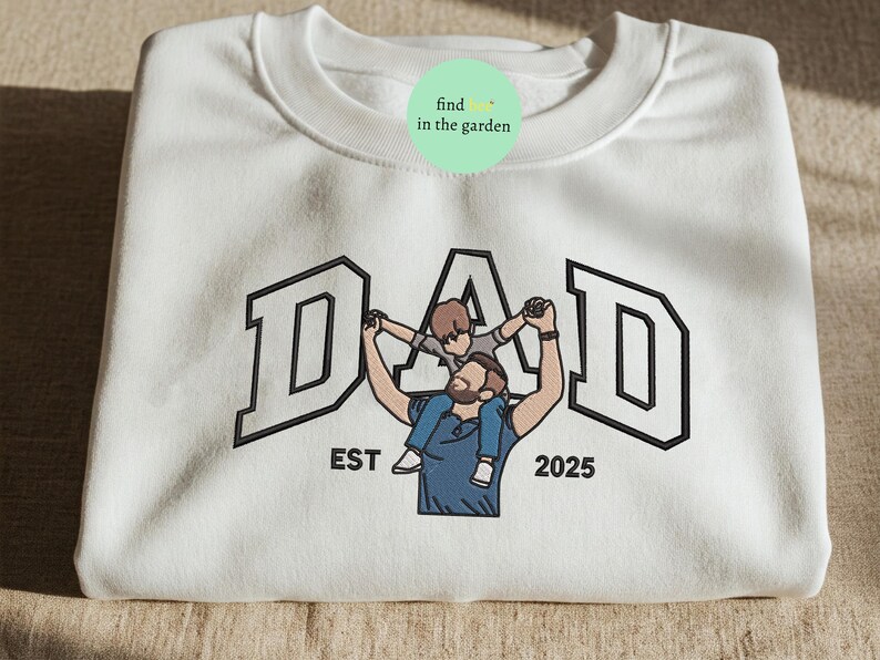 May include: White sweatshirt with the word "DAD" in a black outlined collegiate font. An illustration of a father holding a child on his shoulders is centered below the word. The text "EST 2025" is below the illustration.