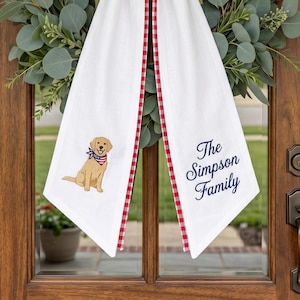 May include: White decorative towel with a red and white checkered trim and a cartoon dog wearing a patriotic bandana. The towel has the text "The Simpson Family" in navy blue script.
