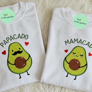 Könnte beinhalten: Zwei weiße Sweatshirts mit Avocado-Designs. Eines trägt die Aufschrift "Papacado" mit einer Avocado mit Schnurrbart und roten Herzen. Das andere trägt die Aufschrift "Mamacado" mit einer lächelnden Avocado und roten Herzen. Beide haben den Text "find in the garden".