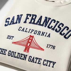May include: Cream-colored sweatshirt with embroidered text and a graphic of the Golden Gate Bridge. The text reads "SAN FRANCISCO CALIFORNIA" above the bridge, and "THE GOLDEN GATE CITY" below. The year "1776" is also included.