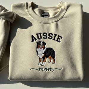 Könnte beinhalten: Cremefarbenes Sweatshirt mit dem schwarzen gestickten Wort "AUSSIE" über einem Bild eines Australian Shepherd Hundes. Unter dem Hund steht das Wort "mom" in grüner Schrift. Das Sweatshirt hat einen Rundhalsausschnitt und lange Ärmel.