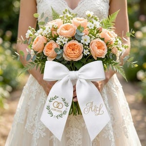 Op de afbeelding: Een bruidsboeket met perzikrozen, witte bloemen en groen. Het boeket is vastgebonden met een groot wit lint. Het lint is geborduurd met gouden initialen en de datum 21.09.2026, samen met een krans en twee in elkaar grijpende ringen.