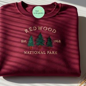 Puede incluir: Sudadera color burdeos con el texto bordado "REDWOOD NATIONAL PARK" y "Est 1968" con tres pinos verdes.