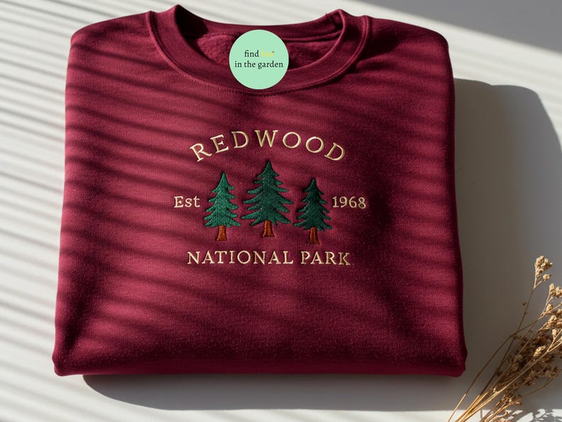 Puede incluir: Sudadera color burdeos con el texto bordado "REDWOOD NATIONAL PARK" y "Est 1968" con tres pinos verdes.