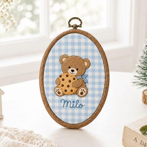 Op de afbeelding: Een ovale houten borduurring met lichtblauw en wit gingham-stof. De stof is voorzien van een geborduurde bruine teddybeer die een beige koekje met chocoladestukjes vasthoudt, met de naam Milo in blauwe draad onder de beer geborduurd.