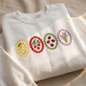 May include: White sweatshirt with four embroidered oval designs. The designs feature a lemon, strawberries, ladybugs, and a bouquet of flowers. Each design has a scalloped border in yellow, red, green, and purple. The sweatshirt is made of a soft, comfortable material.
