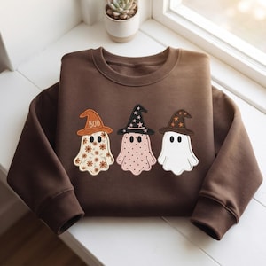 Puede incluir: Una sudadera de cuello redondo color marrón oscuro con un gráfico bordado de tres fantasmas con sombreros de bruja. El fantasma de la izquierda tiene un estampado floral, el del medio lunares y el de la derecha es blanco liso. El sombrero izquierdo tiene el texto BOO.