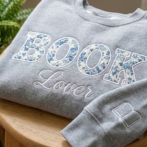 May include: A gray sweatshirt with the words "BOOK Lover" embroidered in white. The letters "BOOK" are filled with a blue floral pattern. A small book graphic is on the sleeve.