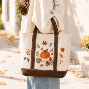 Puede incluir: Un bolso de tela color crema con asas marrón oscuro y una base reforzada. La parte frontal presenta diseños bordados con temática otoñal que incluyen una calabaza naranja grande, una taza verde con malvaviscos, libros y varias hojas.