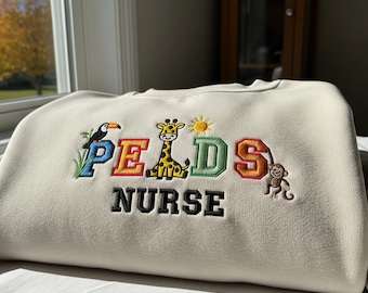 May include: Cream-colored sweatshirt with embroidered text and animal graphics. The word "PEDS" is spelled out in colorful letters, with a toucan, giraffe, and monkey. The word "NURSE" is embroidered in black.
