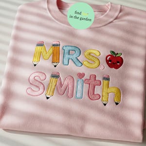 K&ouml;nnte beinhalten: Rosafarbener Pullover mit gesticktem Text "MRS. SMITH" in bunten Buchstaben mit Bleistift- und Apfelmotiven.