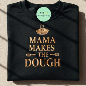 Può includere: Felpa nera con la scritta dorata "MAMA MAKES THE DOUGH". Il design include una grafica a torta e due grafiche a frusta. Un cerchio verde con la scritta "find in the garden" si trova nell'angolo in alto a sinistra.