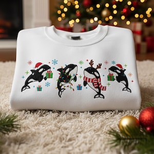 Könnte beinhalten: Weißes Sweatshirt mit einem gestickten Design von vier Orcas, die für Weihnachten geschmückt sind. Die Orcas tragen Weihnachtsmützen, Rentiergeweihe und festliche Schals, mit Schneeflocken und Geschenkpaketen. Ein Weihnachtsbaum ist im Hintergrund zu sehen.
