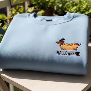 May include: A light blue sweatshirt featuring an embroidered cartoon hot dog wearing a witch hat. The text "Halloweienie" is below the graphic. The sweatshirt is made of a soft material and is perfect for Halloween.