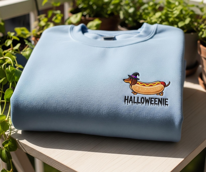 May include: A light blue sweatshirt featuring an embroidered cartoon hot dog wearing a witch hat. The text "Halloweienie" is below the graphic. The sweatshirt is made of a soft material and is perfect for Halloween.