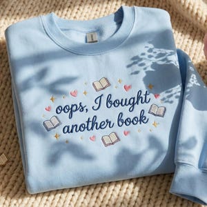 May include: A pale blue sweatshirt embroidered with the words "oops, I bought another book". The design features open books, hearts, and stars. A gold book pendant necklace and a vase with a pink rose are in the background.
