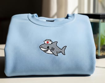 Pode incluir: Camisola azul clara com um tubarão de desenho animado bordado usando um chapéu de enfermeira. O tubarão é cinza com detalhes brancos e uma expressão sorridente. A camisola está dobrada sobre uma superfície branca.