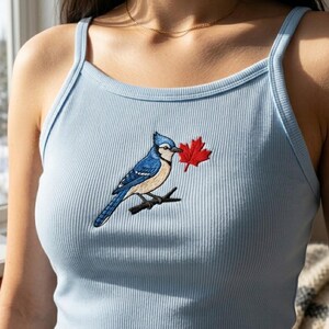 May include: A light blue ribbed tank top featuring an embroidered design of a blue jay perched on a small branch, holding a vibrant red maple leaf in its beak. The garment has thin shoulder straps and a high, rounded neckline.